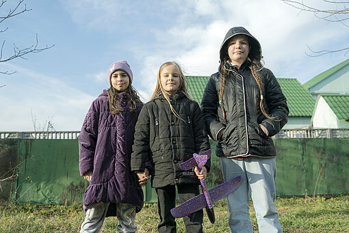 Drei Mädchen stehen auf einer Wiese vor Häusern. Sie tragen dicke Jacken, eine von ihnen hält ein Spielflugzeug in der Hand