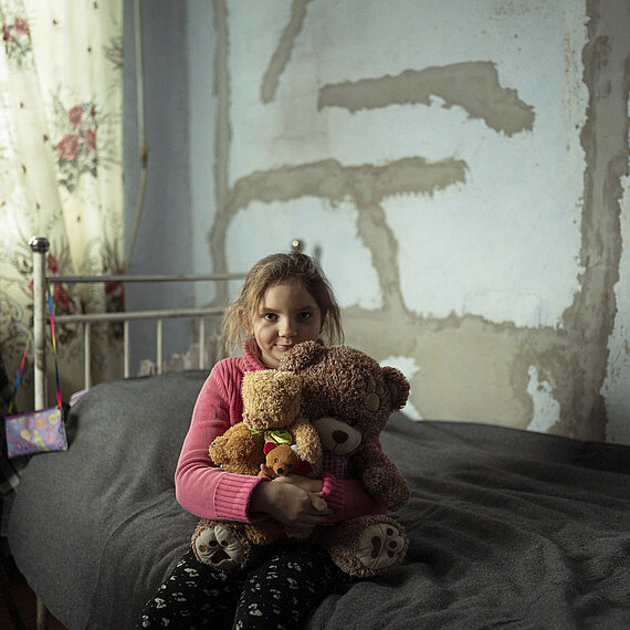 Die 9-jährige Natalka aus der Ukraine sitzt auf einem Bett und hält zwei Teddybären in ihren Armen. Der Raum hat eine unvollständige Wand mit sichtbaren Putzstellen, und es gibt Pflanzen und Vorhänge im Hintergrund.