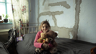 Die 9-jährige Natalka aus der Ukraine sitzt auf einem Bett und hält zwei Teddybären in ihren Armen. Der Raum hat eine unvollständige Wand mit sichtbaren Putzstellen, und es gibt Pflanzen und Vorhänge im Hintergrund.