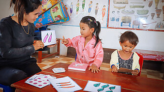 Eine Frau sitzt mit zwei kleinen Kindern an einem Tisch. Sie hält Bilder von Gemüse in der Hand.