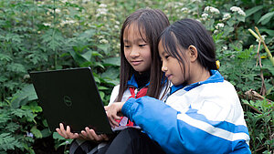 Zwei Mädchen aus Vietnam sitzen in der Natur und lernen gemeinsam am Laptop, umgeben von grünen Pflanzen und Blumen.