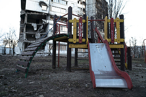 Ein von Bomben zerstörtes Gebäude im Hintergrund, davor ein Kinderspielplatz