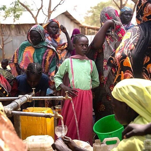 Ein Mädchen steht in einer Gruppe von Menschen, die sich um einen Wasserspender versammelt hat.