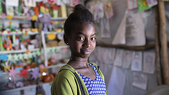 Ein Mädchen steht in einem Zelt und schaut in die Ferne. Im Hintergrund hängen bunte Bilder, von Kindern gemalt.