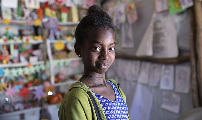 Ein Mädchen steht in einem Zelt und schaut in die Ferne. Im Hintergrund hängen bunte Bilder, von Kindern gemalt.