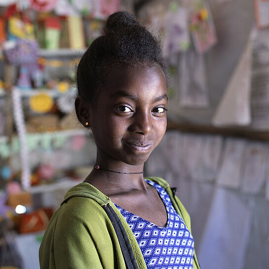 Ein Mädchen steht in einem Zelt und schaut in die Ferne. Im Hintergrund hängen bunte Bilder, von Kindern gemalt.