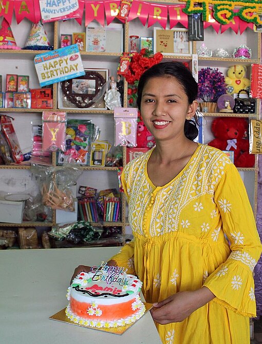 Basanti steht in einem gelben Kleid vor einem Regal mit ihren Produkten und hält lächelnd eine Torte in den Händen