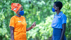 Eine Plan-Mitarbeiterin im orangenen Shirt spricht mit einer Schülerin in Schuluniform – beide tragen Masken zum Schutz vor Corona.