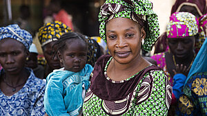 Selbstbewusste Frau in traditioneller Kleidung hält ein Kind im Arm, umgeben von weiteren Frauen in bunter afrikanischer Kleidung