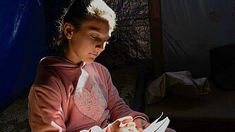 Ein Mädchen sitzt in einem dunklen Zelt und malt ein Bild, ein einzelner Lichtstrahl fällt auf ihr Gesicht