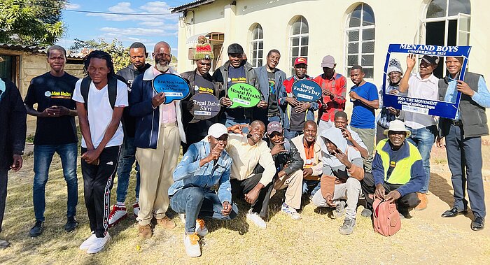 Eine Gruppe von Männern posiert für ein Foto vor einer gelben Wand mit Fenstern. Sie halten Schilder mit Botschaften, teilweise zum Thema Männergesundheit und Gemeinschaft.