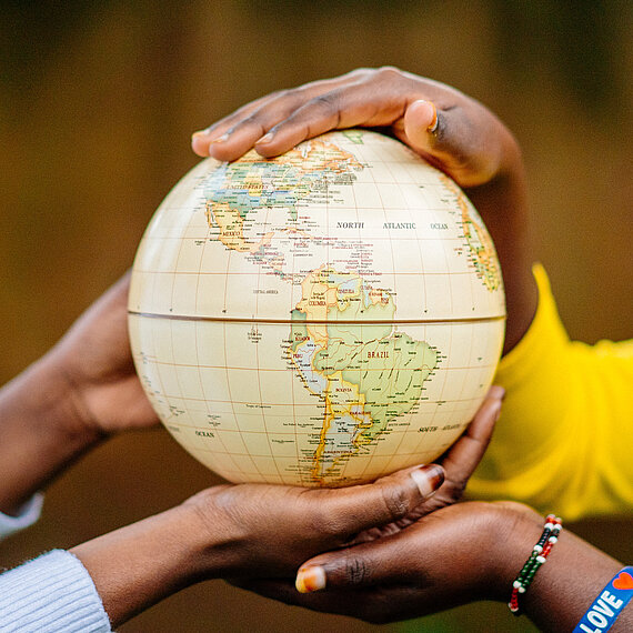 Mehrere Hände – vermutlich von jungen Frauen – halten gemeinsam eine Weltkugel, die auf Nord- und Südamerika ausgerichtet ist.