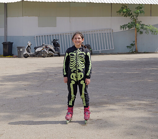 Ein Kind trägt Rollschuhe und steht draußen in einem Innenhof. Es hat einen schwarzen Trainingsanzug mit leuchtendem Skelettmotiv an. Gebäude und geparkte Motorroller sind im Hintergrund sichtbar.