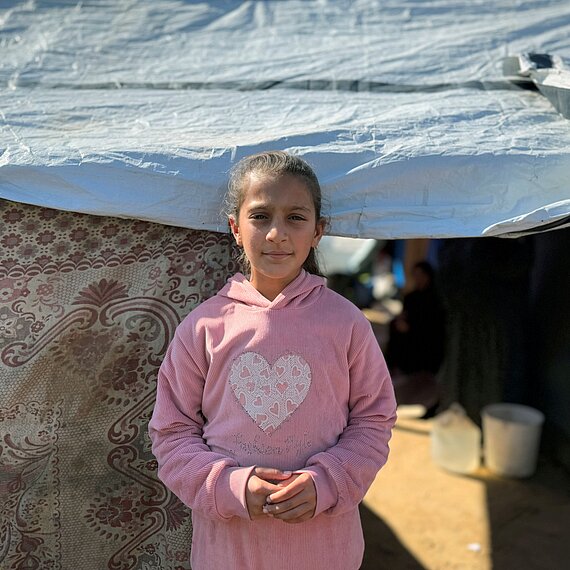 Ein Mädchen steht vor einem provisorischen Zelt, das mit einer Plane und dekorativen Stoffen abgedeckt ist. Das Kind trägt einen rosa Kapuzenpullover mit einem großen Herz auf der Brust. Im Hintergrund sieht man einen sandigen Boden, einen weißen Eimer und im Halbschatten weitere Personen sowie den Eingang zum Zelt.
