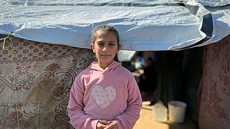 Ein Mädchen steht vor einem provisorischen Zelt, das mit einer Plane und dekorativen Stoffen abgedeckt ist. Das Kind trägt einen rosa Kapuzenpullover mit einem großen Herz auf der Brust. Im Hintergrund sieht man einen sandigen Boden, einen weißen Eimer und im Halbschatten weitere Personen sowie den Eingang zum Zelt.