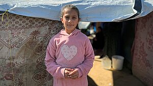 Ein Mädchen steht vor einem provisorischen Zelt, das mit einer Plane und dekorativen Stoffen abgedeckt ist. Das Kind trägt einen rosa Kapuzenpullover mit einem großen Herz auf der Brust. Im Hintergrund sieht man einen sandigen Boden, einen weißen Eimer und im Halbschatten weitere Personen sowie den Eingang zum Zelt.