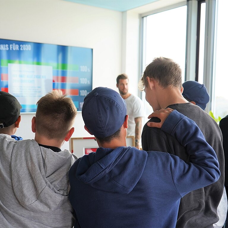 Eine Gruppe Schüler steht im Halbkreis vor einem großen Bildschirm. Man sieht die Schüler von hinten.