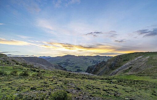 Panorama-Blick über Wiesen, Hügel und Berge, über denen die Sonne aufgeht