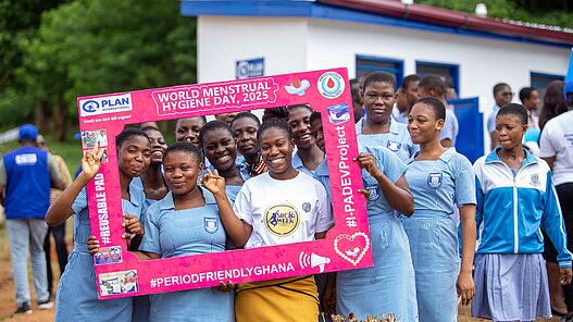 Mehrere Mädchen in Schuluniform stehen beieinander und lachen durch einen pinken Rahmen in die Kamera. Der Rahmen macht auf den World Menstrual Hygiene Day aufmerksam.