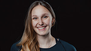 Magdalena Matschine steht vor einem schwarzen Hintergrund, trägt ihre Haare über die rechte Schulter und lächelt in die Kamera.