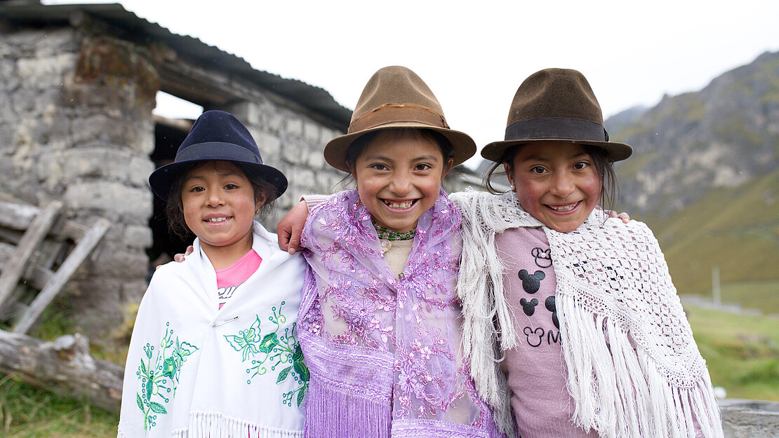 Drei Mädchen mit Hüten und Ponchos stehen auf einer Wiese, haben die Arme um die Schultern gelegt und lächeln in die Kamera