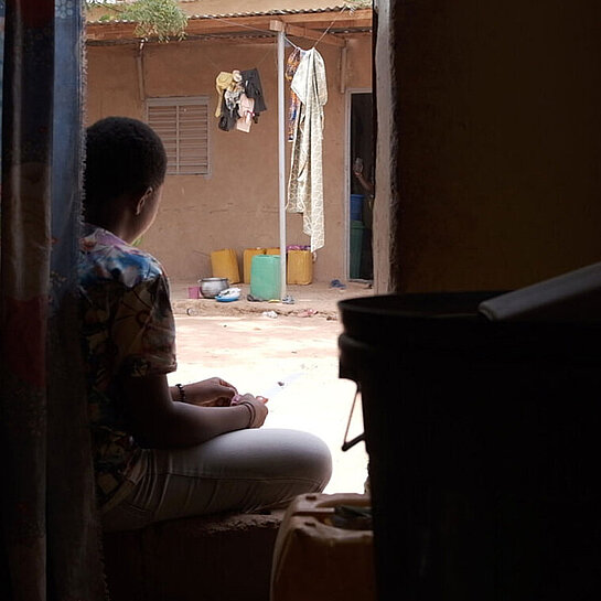 Durch einen Hauseingang sieht man eine junge Frau vor der Tür im Hof sitzen.
