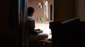 Durch einen Hauseingang sieht man eine junge Frau vor der Tür im Hof sitzen.