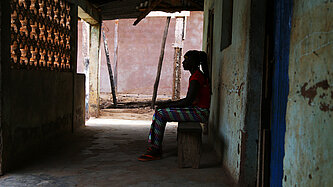 Eine Frau sitzt vor einem Haus unter einem Vordach, man sieht nur ihre schwarze Silhouette 