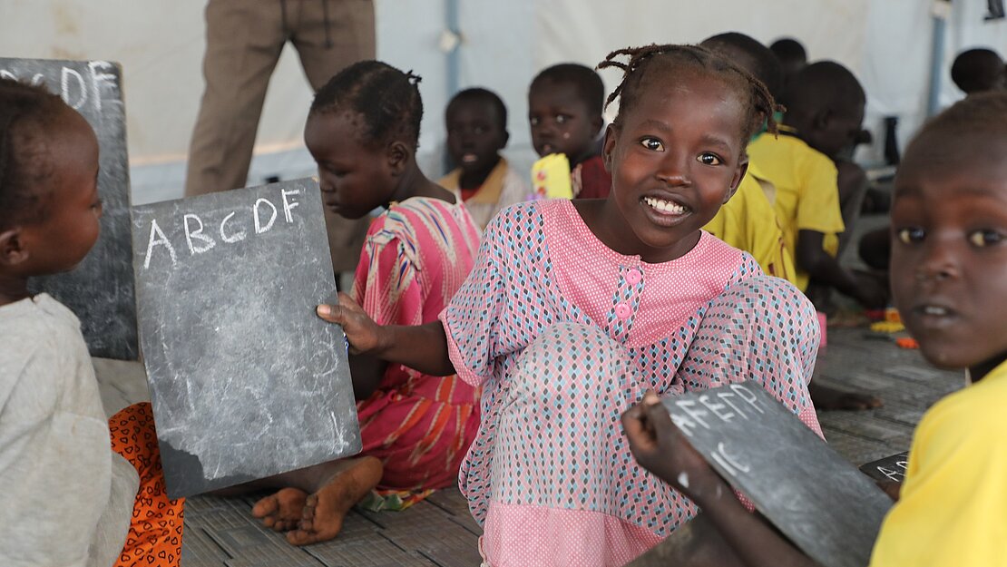 Ein Mädchen hält lächelnd eine Schiefertafel, auf der die Buchstaben A, B, C, D, und F stehen