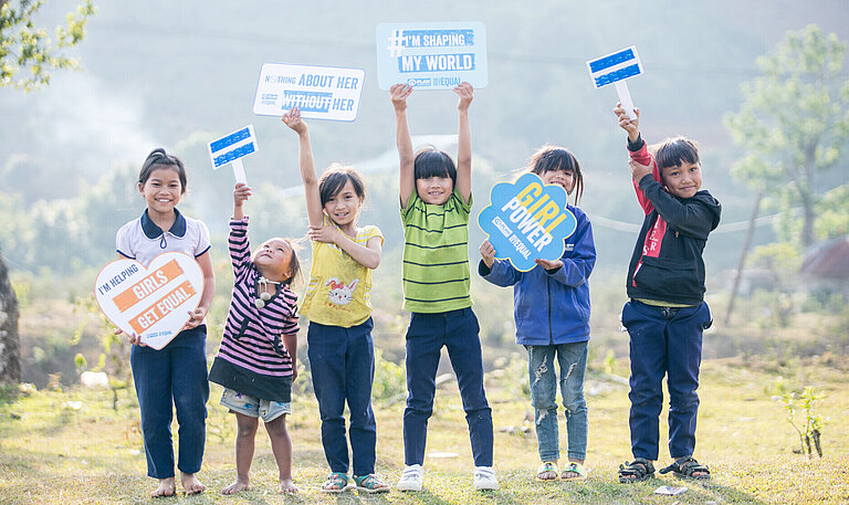 Eine Gruppe von Kindern in Vietnam hält Schilder mit Botschaften wie "I'm helping girls get equal" und "Girl power". Sie stehen auf einer Wiese und zeigen ihre Unterstützung für Geschlechtergleichstellung.
