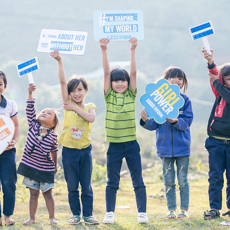 Eine Gruppe von Kindern in Vietnam hält Schilder mit Botschaften wie "I'm helping girls get equal" und "Girl power". Sie stehen auf einer Wiese und zeigen ihre Unterstützung für Geschlechtergleichstellung.