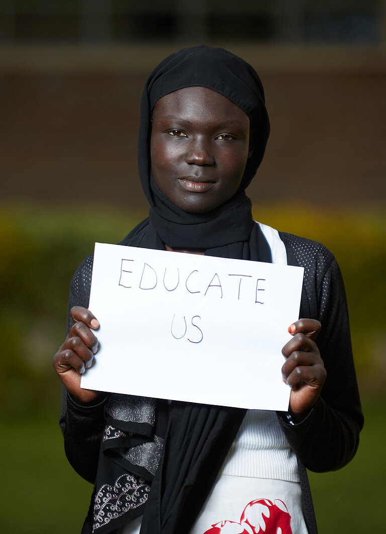 Ein Mädchen hält ein Schild hoch, auf dem Educate Us (bildet uns) steht