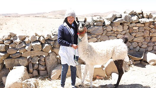 Eine Frau steht neben einem Lama und hält es an der Leine. Sie grinst in die Kamera. 