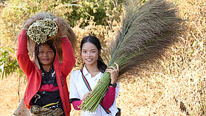 Zwei Frauen in Laos tragen frisch geerntetes Besengras – eine trägt ein Bündel auf dem Kopf, die andere hält ein großes Bündel in der Hand.