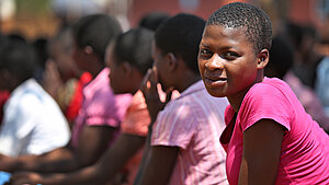Ein junges Mädchen in Malawi trägt ein pinkes T-Shirt und lächelt in die Kamera, während sie bei einer Versammlung im Freien sitzt. Im Hintergrund sind weitere Jugendliche in bunter Kleidung zu sehen.