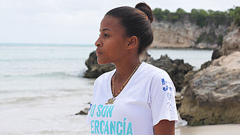 Ein Mädchen mit Dutt, im Profil zu sehen, steht an einem Strand. Im Hintergrund sieht man Meer und Klippen.
