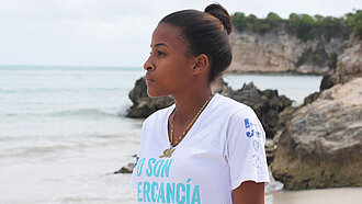 Ein Mädchen mit Dutt, im Profil zu sehen, steht an einem Strand. Im Hintergrund sieht man Meer und Klippen.