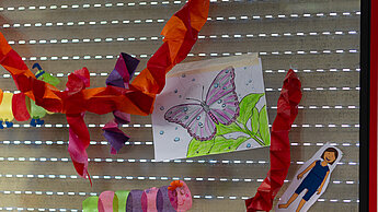Bunte Kinderkunstwerke, darunter ein gemalter Schmetterling und aus Papier gestaltete Raupen, hängen an einem Fenster.