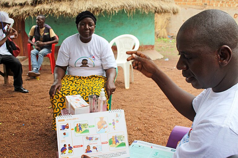 Ein Mann hält ein Faktenblatt mit Symbolen in der Hand und erklärt einer Frau, die ihm gegenüber sitzt, etwas über Malaria