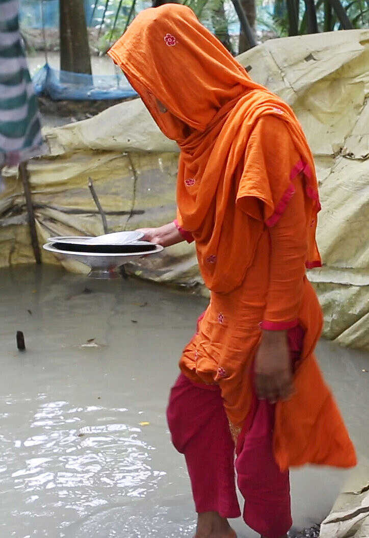 Keya steht neben einem Gewässer und hält Geschirr in der Hand. Sie trägt ein leuchtend orangefarbenes Gewand.