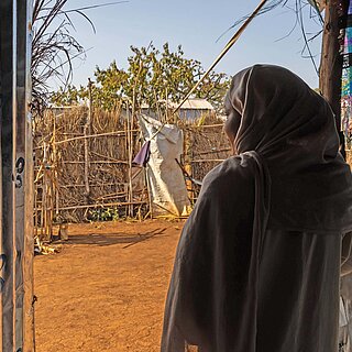 Eine Frau mit Hijab steht in einer provisorischen Unterkunft in Ura. Sie steht im Schatten und blickt auf die sonnige Umgebung vor ihr mit weiteren Unterkünften.