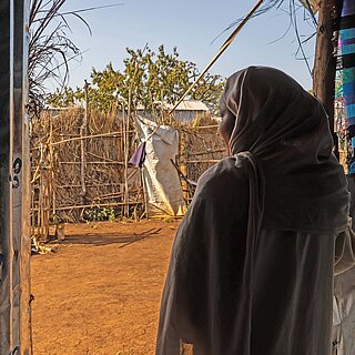 Eine Frau mit Hijab steht in einer provisorischen Unterkunft in Ura. Sie steht im Schatten und blickt auf die sonnige Umgebung vor ihr mit weiteren Unterkünften.