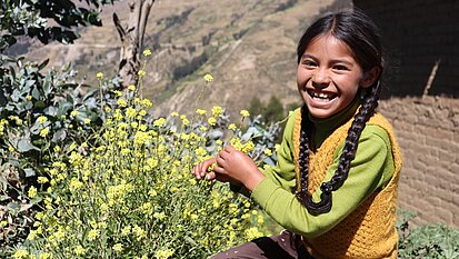 Ein Mädchen sitzt vor einer Berglandschaft in Peru. Sie lacht freudig und hält den Stiel einer gelben Blume in der Hand.