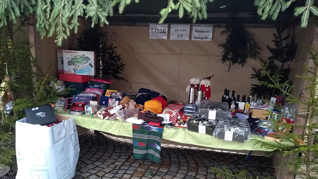 Tombola-Stand mit diversen Preisen wie Stofftieren, Büchern, Thermoskannen und Getränken auf einem Weihnachtsmarkt zugunsten der Schönebeck-Stiftung