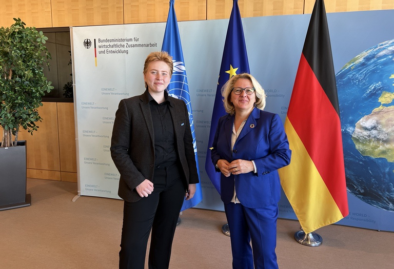 Henrike und Svenja Schulze stehen vor drei Flaggen: UN, Europa und Deutschland. Beide tragen Anzüge und lächeln in die Kamera.