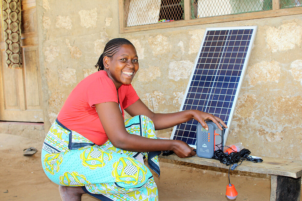 Frau in Kenia zeigt ein tragbares Solarsystem vor ihrem Haus als Teil eines Projekts für nachhaltige Energieversorgung