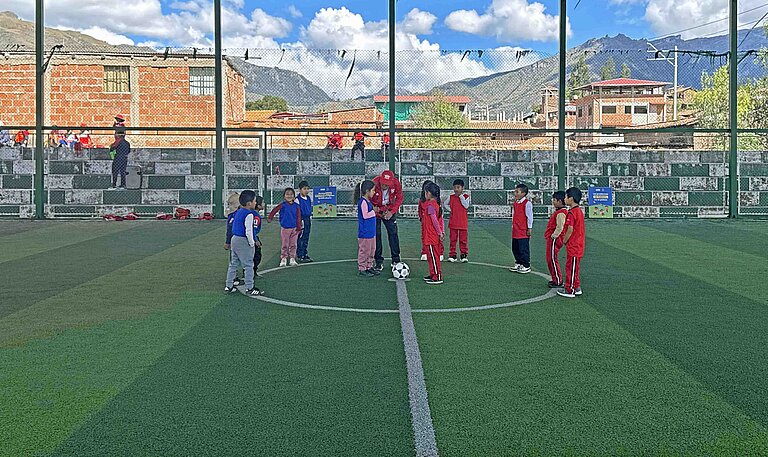 Mädchen und Jungen stehen in Trikots auf einem überdachten Fußball-Platz, im Hintergrund sind schneebedeckte Berge sichtbar.