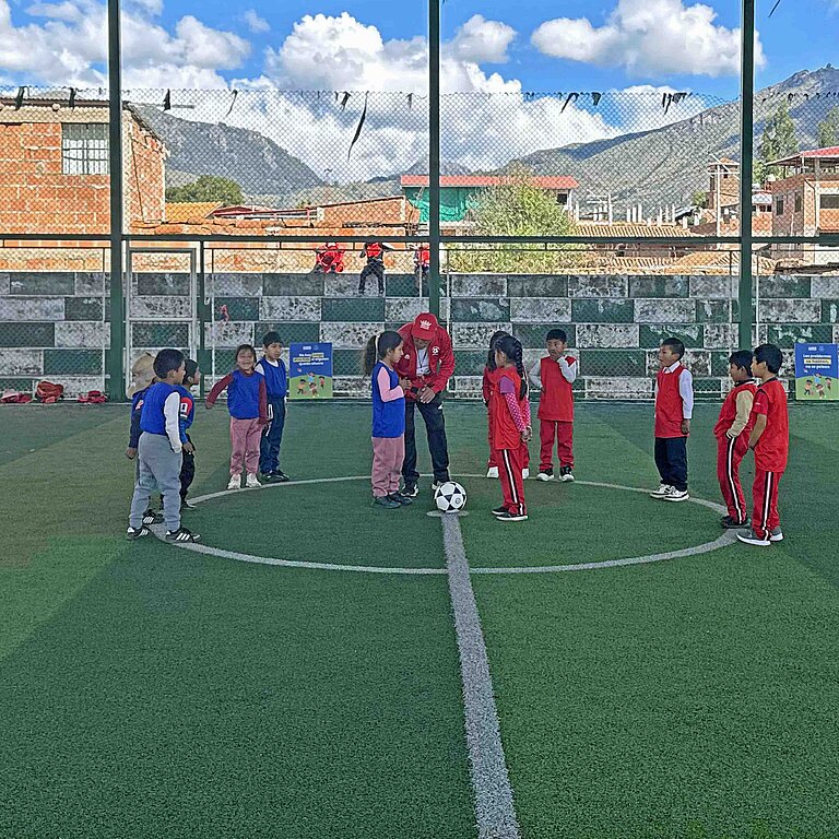 Mädchen und Jungen stehen in Trikots auf einem überdachten Fußball-Platz, im Hintergrund sind schneebedeckte Berge sichtbar.