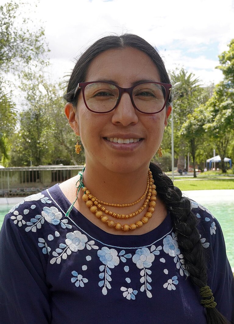 Verónica steht im Freien vor einer Wasserquelle in einer ländlichen Umgebung und blickt in die Kamera