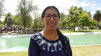 Verónica steht im Freien vor einer Wasserquelle in einer ländlichen Umgebung und blickt in die Kamera